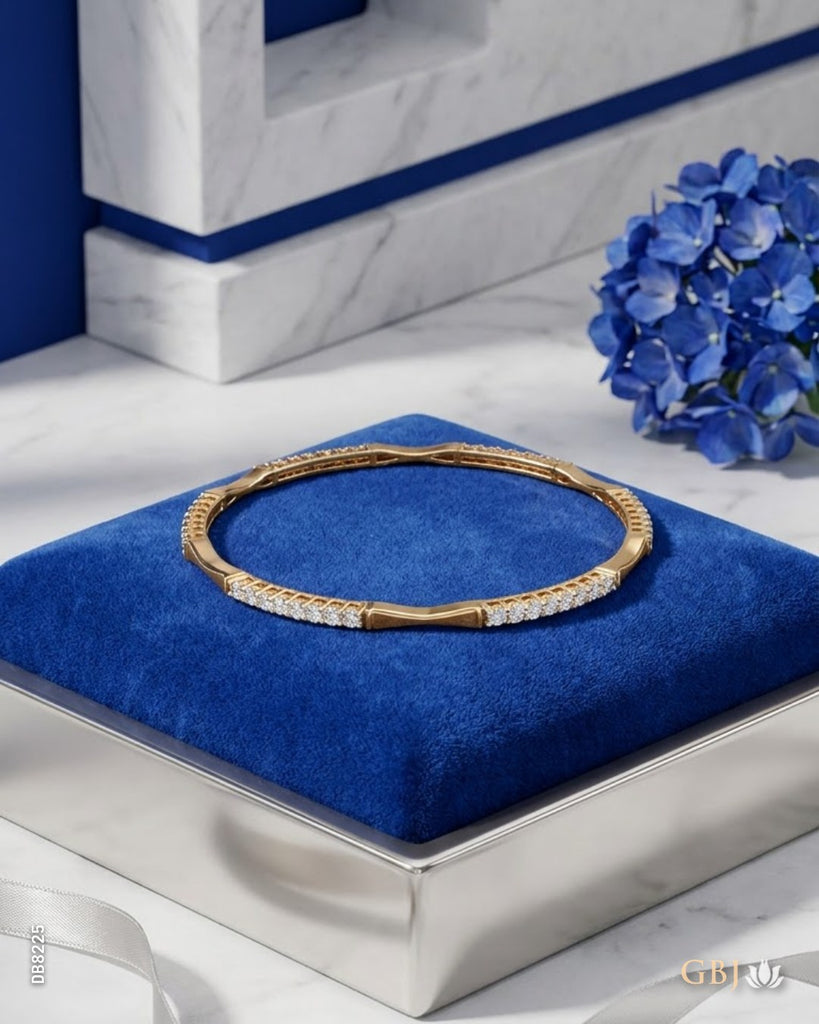 A gold bangle bracelet with a row of clear stones, displayed on a blue velvet jewelry box on a marble surface with blue flowers in the background.