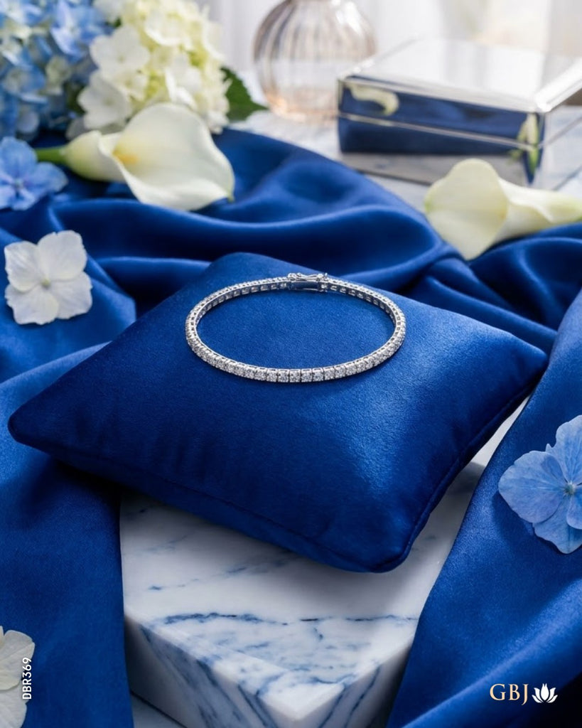 A silver tennis bracelet set with clear stones, displayed on a royal blue velvet cushion. The cushion is placed on a white marble block, surrounded by draped royal blue satin fabric, white and blue flowers, and a small blue jewelry box in the background.