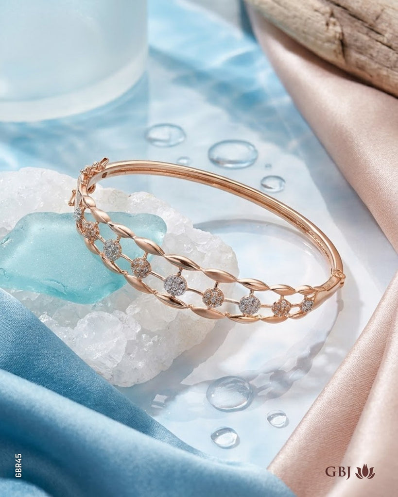 A rose gold bangle bracelet with a twisted chain design and small clear gemstone accents, styled on white crystals with blue and pink satin fabrics.