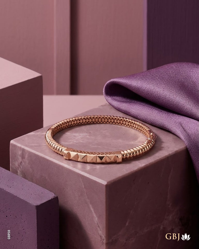 A rose gold bangle bracelet with a textured honeycomb pattern and a pyramid-link closure, displayed on a purple marble block with purple fabric.