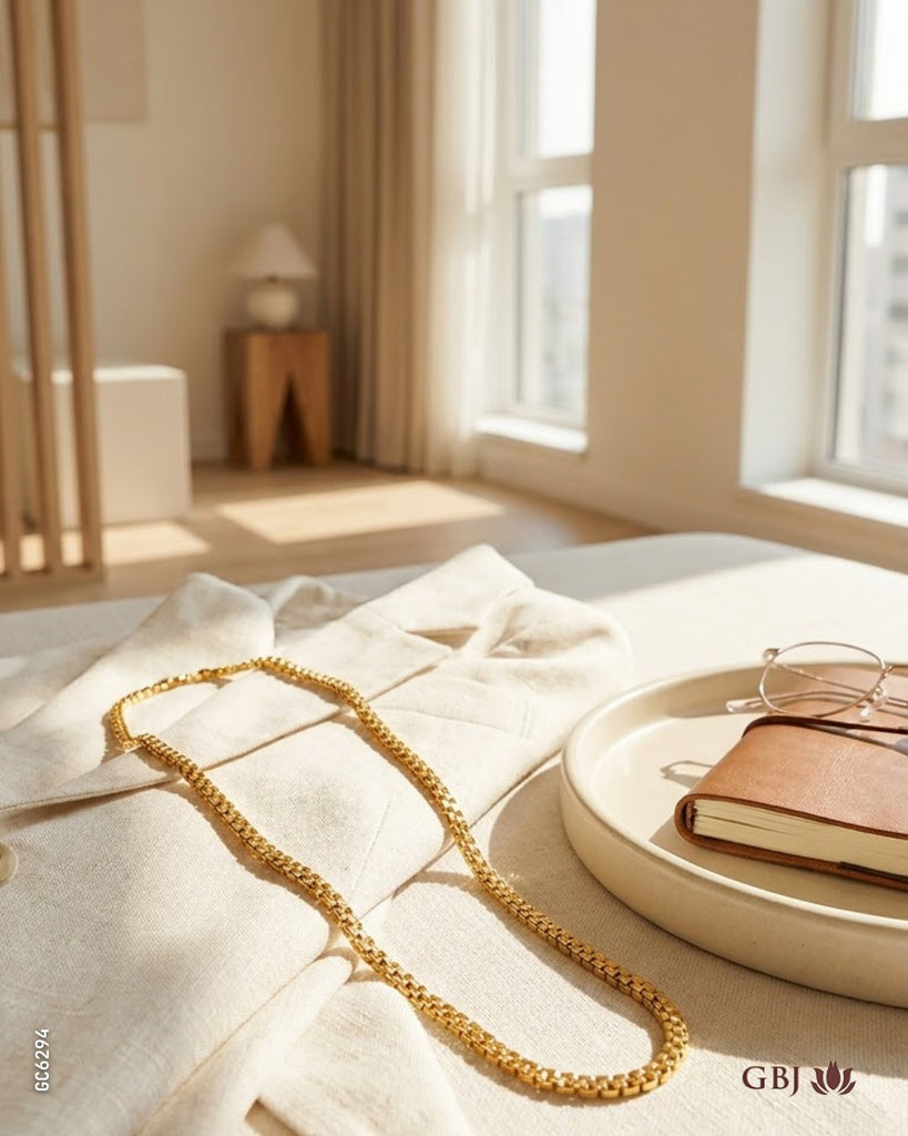 A gold chain necklace draped elegantly over a light beige linen fabric, styled on a bed in a bright, sunlit bedroom. A small brown notebook and eyeglasses rest on a round tray nearby.
