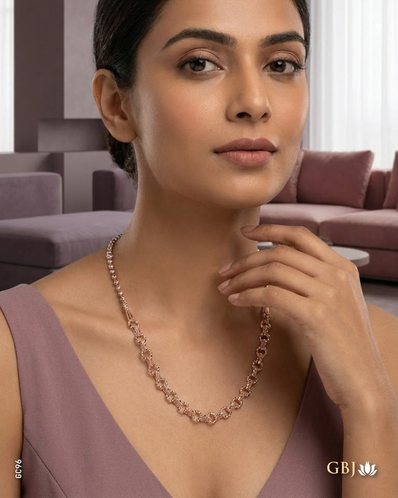 A woman wearing a delicate rose gold chain necklace with small sparkling stones, posed in a modern living room setting. The brand logo 'GBJ' is visible in the bottom right corner.