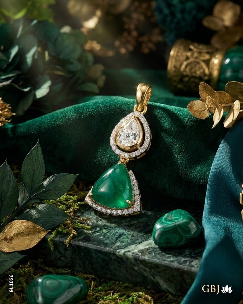 A gold pendant necklace with a large green teardrop-shaped stone and a smaller clear teardrop stone above it, styled on a dark green velvet cloth with green stones and gold leaves.