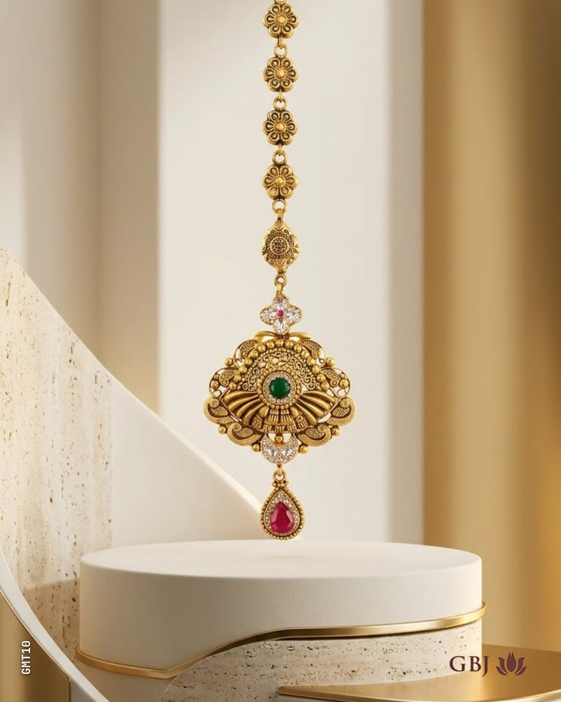 An ornate gold-toned traditional Indian jewelry piece with a long chain, a large fan-shaped pendant featuring a green stone, and a teardrop-shaped red stone, displayed on a white pedestal.
