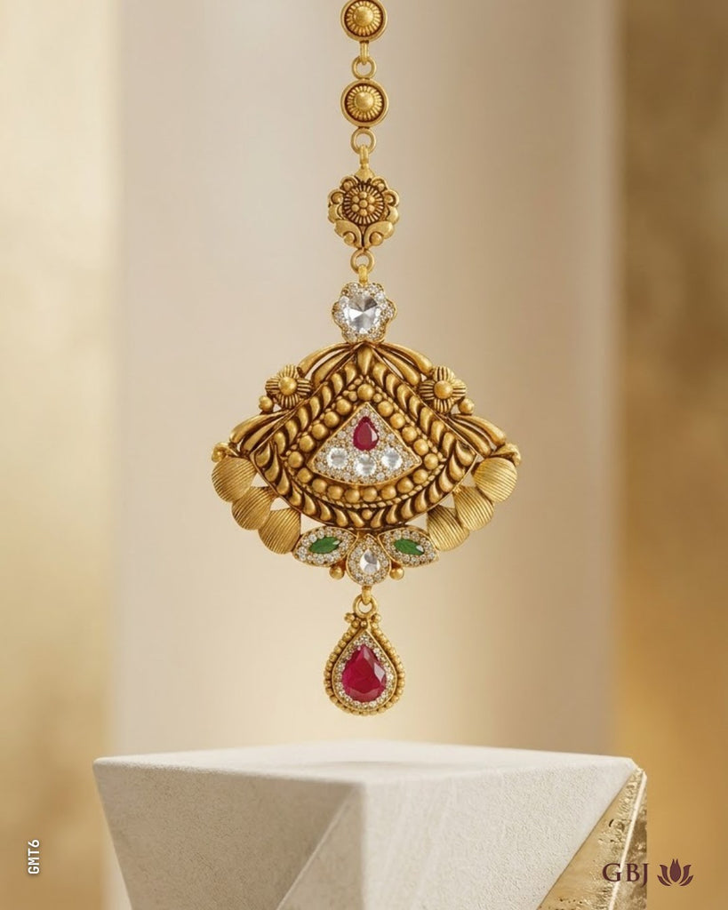 An ornate gold-toned traditional Indian jewelry piece, likely a pendant or part of a necklace, displayed on a white pedestal. The piece features intricate gold filigree, a central clear heart-shaped stone, a small red stone, green stones, and a teardrop-shaped red stone at the bottom.