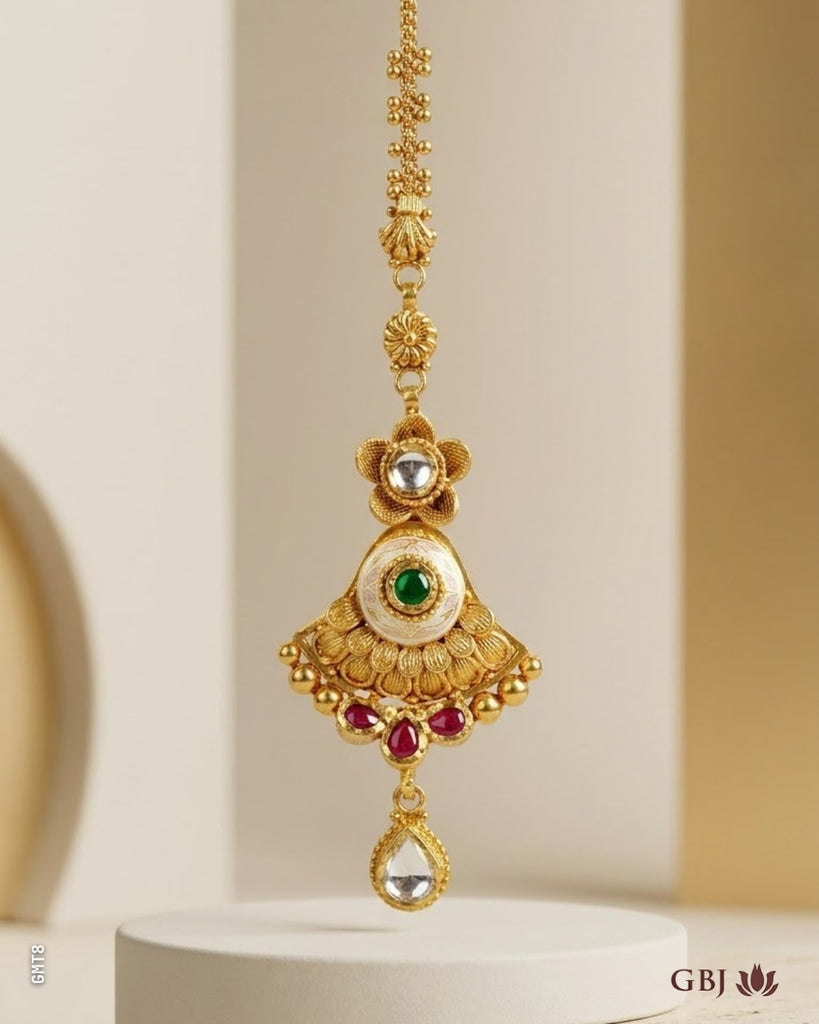 An ornate gold-toned traditional pendant necklace with a teardrop-shaped pendant, featuring a central green stone, red accent stones, and a clear teardrop gemstone, displayed on a white pedestal.
